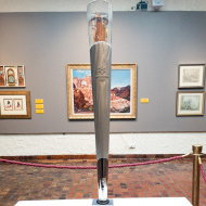 photo of Olympic Torch on a white pedestal with several paintings in the background