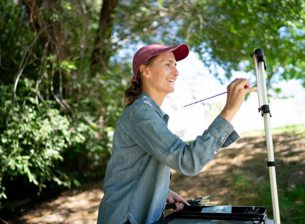 smiling adult woman with hat painting outdoors surrounded by trees