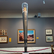 photo of Olympic Torch on a white pedestal with several paintings in the background